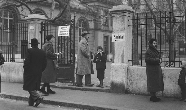 Verschiedene Menschen mit Wahlcouvert in der Hand betreten durch ein Tor den Hof eines Schulhauses und Wahllokals.